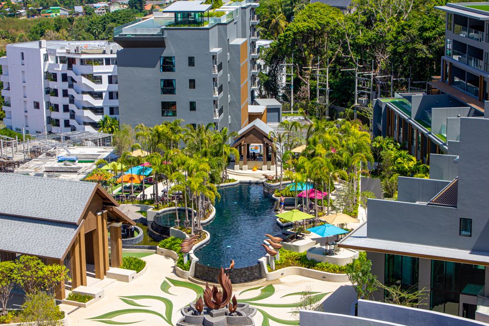 The image depicts a vibrant outdoor area featuring a large swimming pool surrounded by green palms and colorful lounge chairs. Multiple buildings create a modern ambiance, with shaded relaxation spots and pathways. Sunlight creates a warm atmosphere, enhancing the inviting nature of the space.