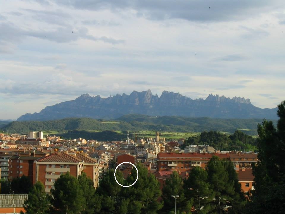 A panoramic view showcases the surrounding landscape of Manresa, characterized by a mixture of urban development and natural scenery. The iconic mountain range in the background is framed by soft clouds and a gentle sky, while trees in the foreground provide a sense of greenery and depth.