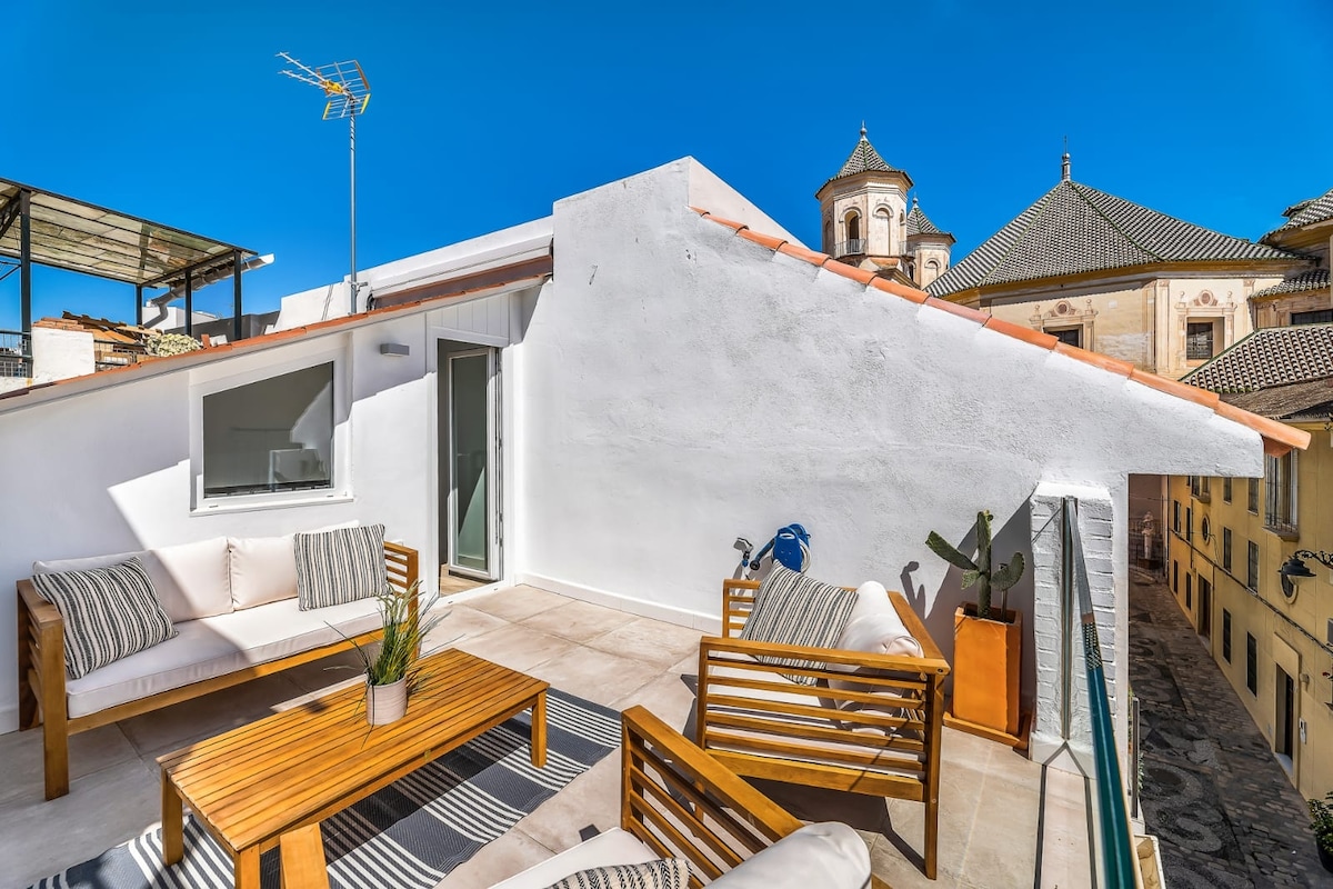 A sunlit rooftop terrace features wooden seating and a coffee table, surrounded by views of historic buildings. The space is designed for relaxation, with a clear sky overhead. A small plant is positioned in a corner, adding a touch of greenery to the setting.