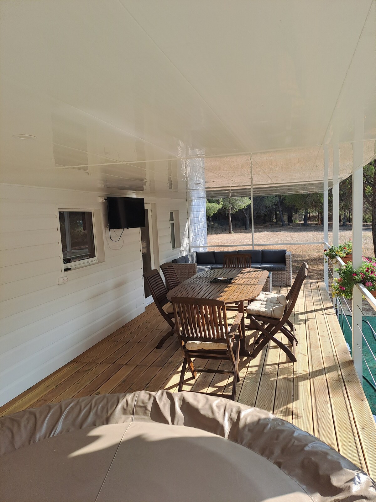 An outdoor terrace features a wooden dining table surrounded by matching chairs, with a comfortable seating area nearby. A Jacuzzi is positioned to the side, and a flat-screen television is mounted on the wall. Natural surroundings are visible in the background.