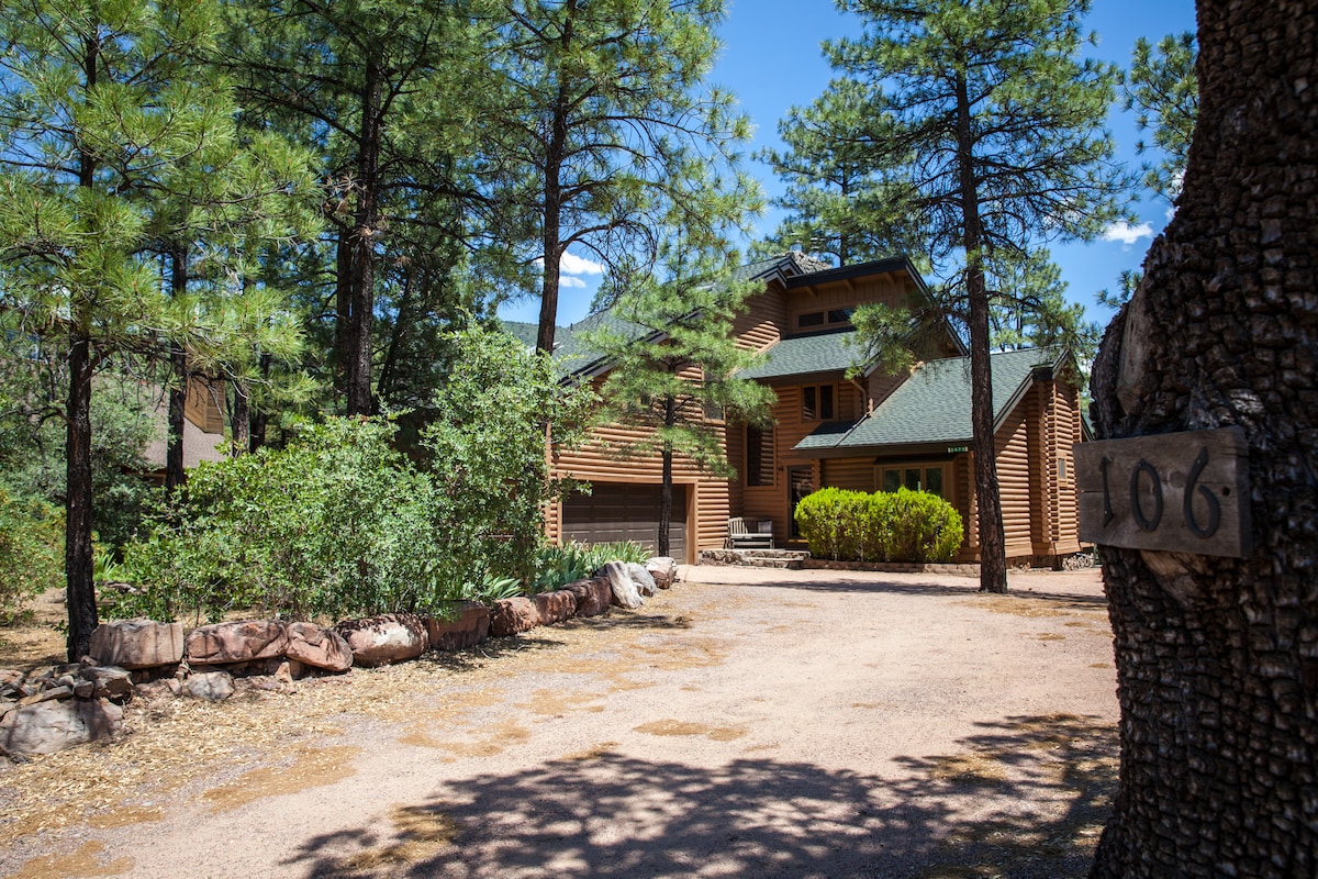 The exterior of the mountain cabin is surrounded by tall pine trees, showcasing a natural setting. A gravel driveway leads to the home, which features wooden siding and a green roof. The entrance is marked by a rustic house number plaque affixed to a nearby tree.