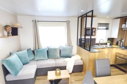 A comfortable living area is shown, featuring a corner sofa adorned with blue cushions. A modern kitchen with wooden cabinetry and a dining area is also visible. Natural light enters through a window framed by light curtains, creating a cozy ambiance.