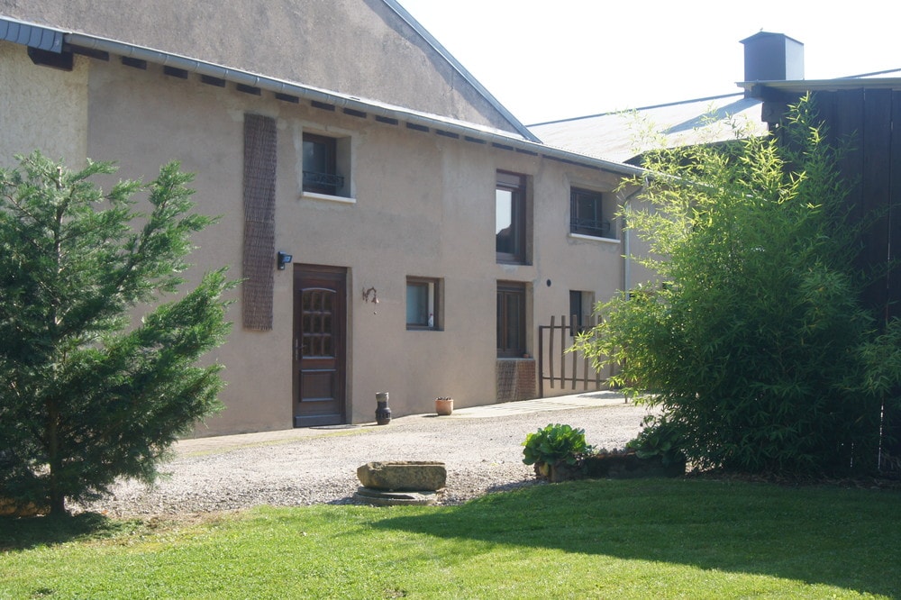 The exterior of the renovated farmhouse is showcased, featuring a combination of natural stone and smooth surfaces. A gravel pathway leads up to a welcoming wooden door framed by windows. Lush greenery and a stone planter enhance the entrance, complementing the rustic charm of the building.