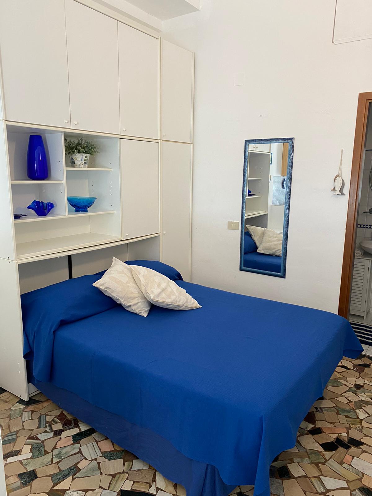 A cozy sleeping area features a blue bedspread draped over a spacious bed, accompanied by decorative pillows. Wall-mounted shelves hold blue and clear glassware, while a full-length mirror reflects the light and adds depth to the room.