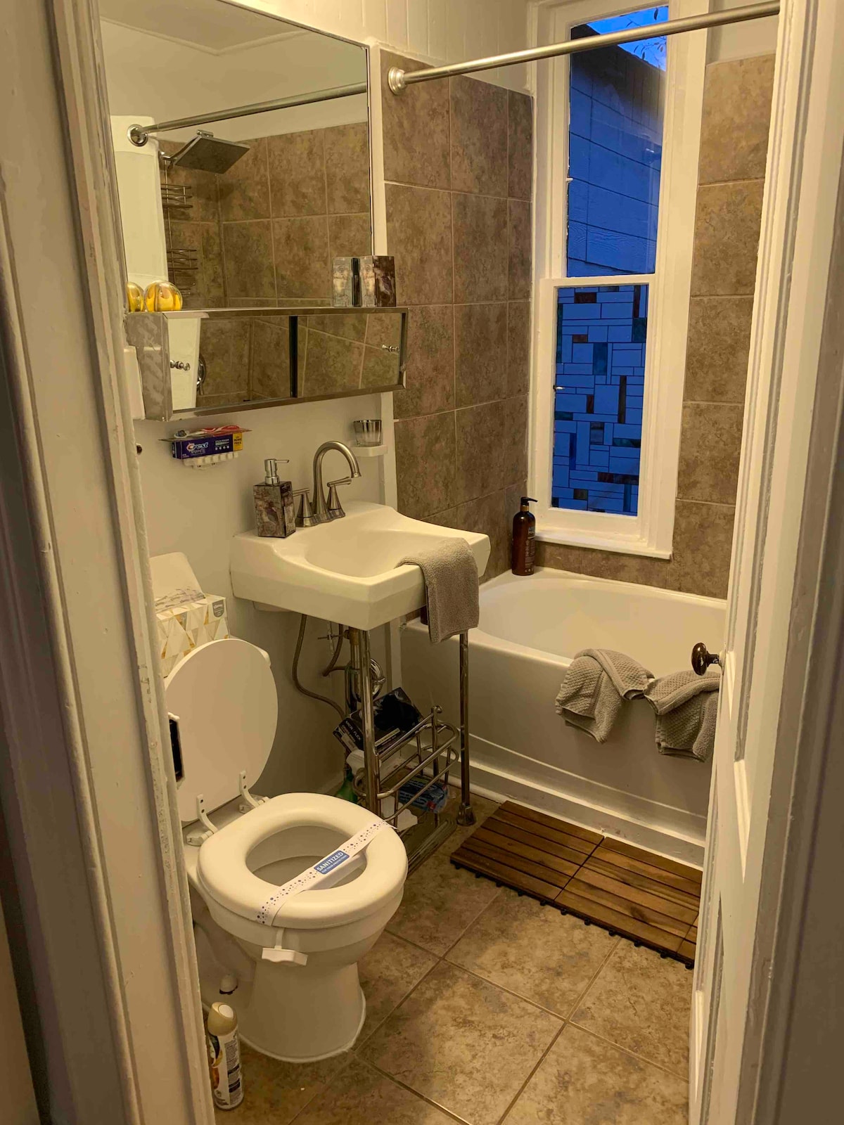 A compact bathroom features a combination tub and shower, with a modern sink mounted on a wall. The area is lined with neutral-toned tiles, and a wooden mat is placed in front of the tub. Natural light enters through a tall window beside the tub.