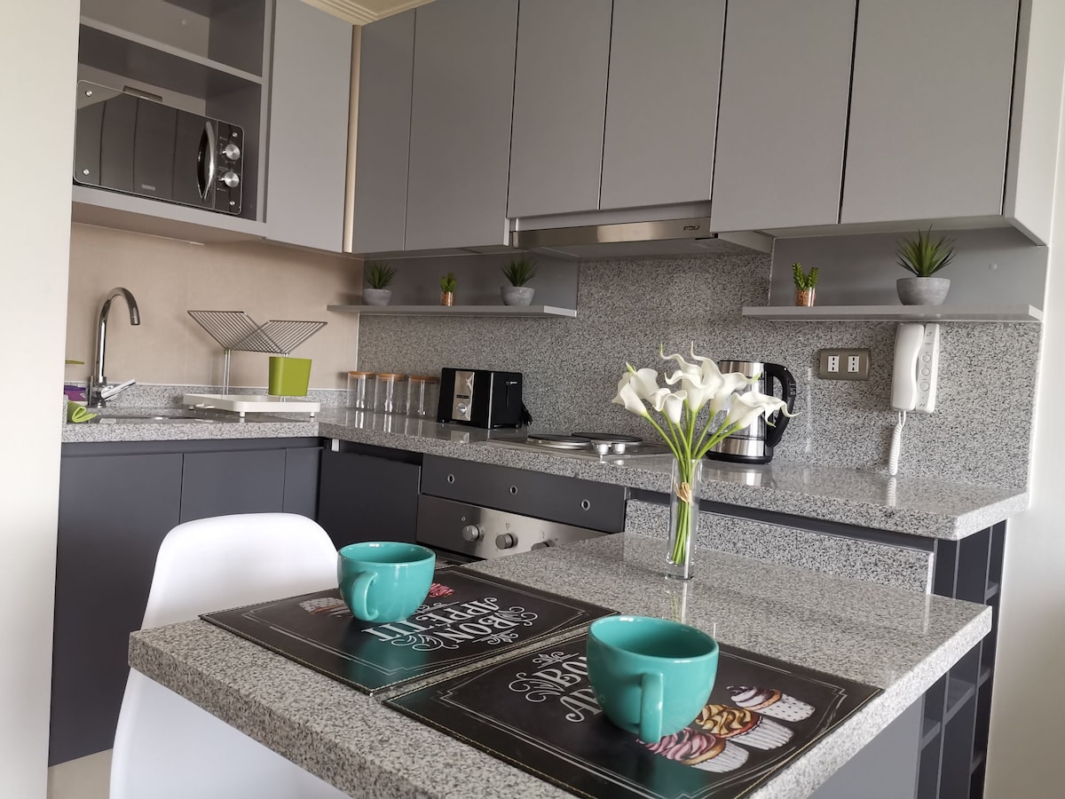 A modern kitchen showcases sleek cabinetry and grey countertops, featuring a coffee maker, toaster, and microwave. The dining area includes a small table set with two teal cups and decorative plants, creating a functional and inviting culinary space.