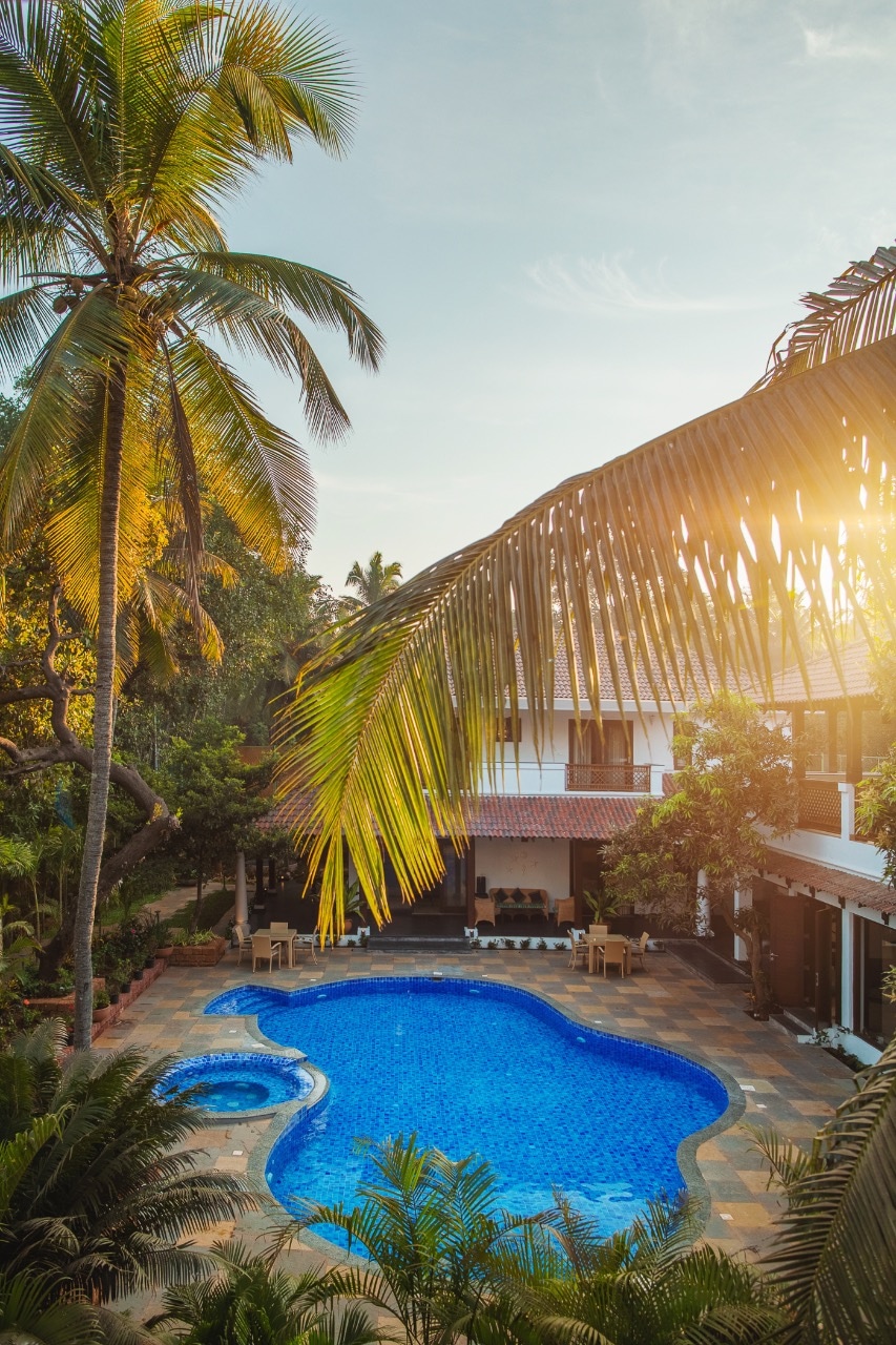 A spacious swimming pool glimmers in turquoise, surrounded by lush greenery and palm trees. Sunlight filters through the leaves, casting warm hues across the area. The charming layout includes inviting outdoor seating near the pool, creating a serene tropical atmosphere.