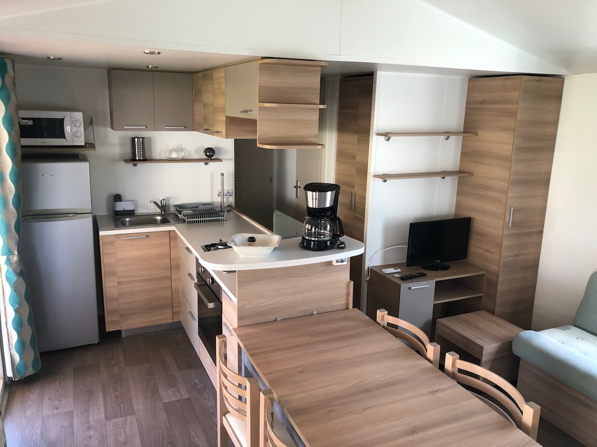 A modern kitchen and dining area are presented, featuring a compact layout with light wood cabinetry. Dining chairs surround a wooden table, while appliances, including a refrigerator and coffee maker, are visible. Natural light enters through a nearby window, creating a bright and functional space.