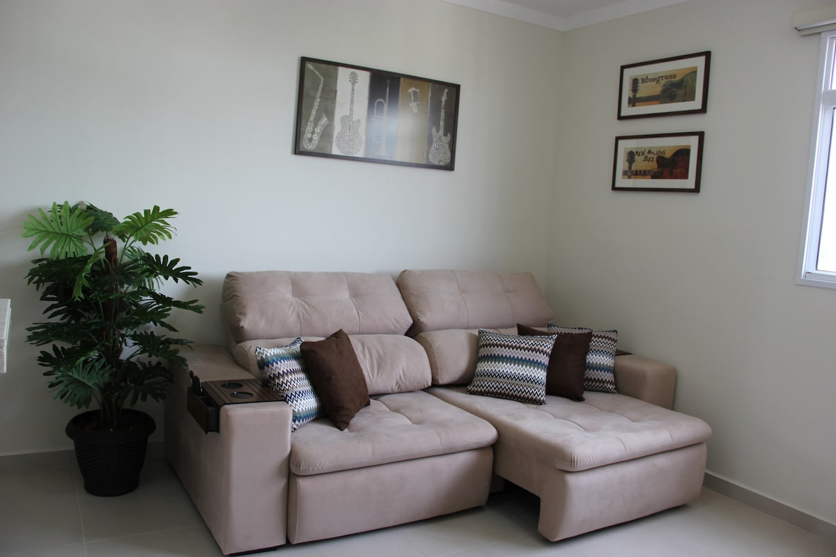 A modern living area features a spacious sectional sofa in light beige, complemented by decorative cushions in neutral tones. A small potted plant adds a touch of greenery, while framed artwork hangs on the wall above, showcasing musical instruments.