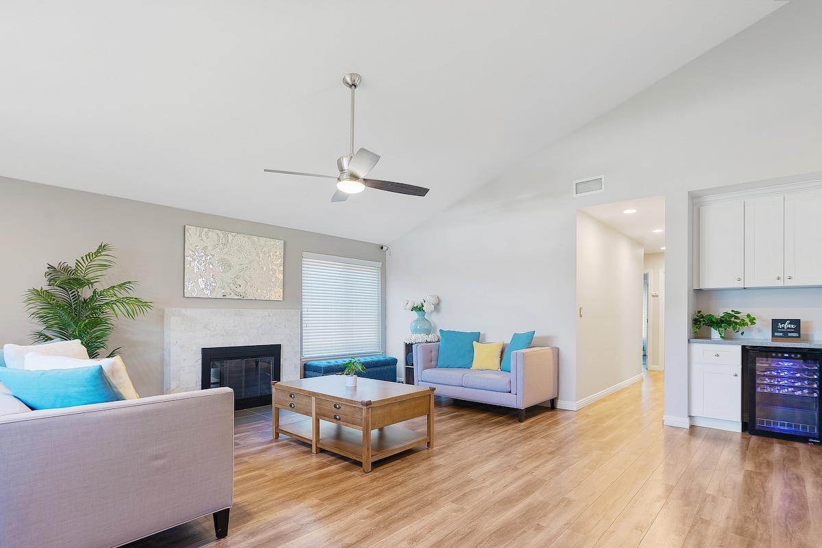 The living room features a spacious layout with vaulted ceilings, two comfortable sofas, and a coffee table. A decorative mirror hangs above a modern fireplace, while a wine and beverage fridge adds convenience. Natural light enters through the window, enhancing the inviting ambiance.