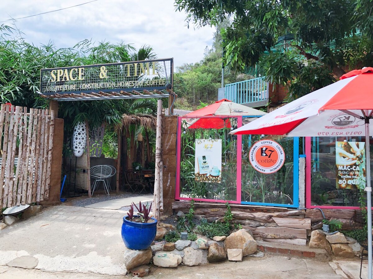 An outdoor area is presented with a wooden gate leading into a vibrant coffee space. Colorful umbrellas provide shade for seating areas. Lush greenery surrounds the entrance, complemented by decorative elements like stones and plants, creating an inviting setting for guests.