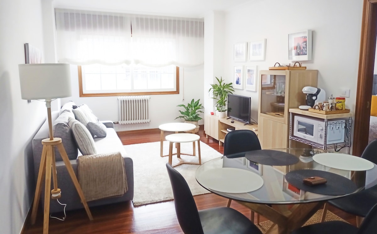 A light-filled living area features a comfortable gray sofa, accompanied by two tables and an armchair. A decorative plant adds a touch of greenery. A dining table is set for six, and a television is positioned on a wooden cabinet, enhancing the inviting space.