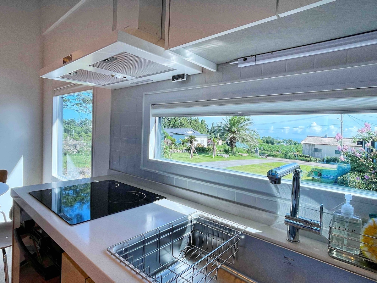 A modern kitchen is equipped with a sleek countertop and a stainless-steel sink. A large window overlooks a landscaped area, allowing natural light to fill the space. The stove is situated on the counter nearby, contributing to a functional cooking environment.