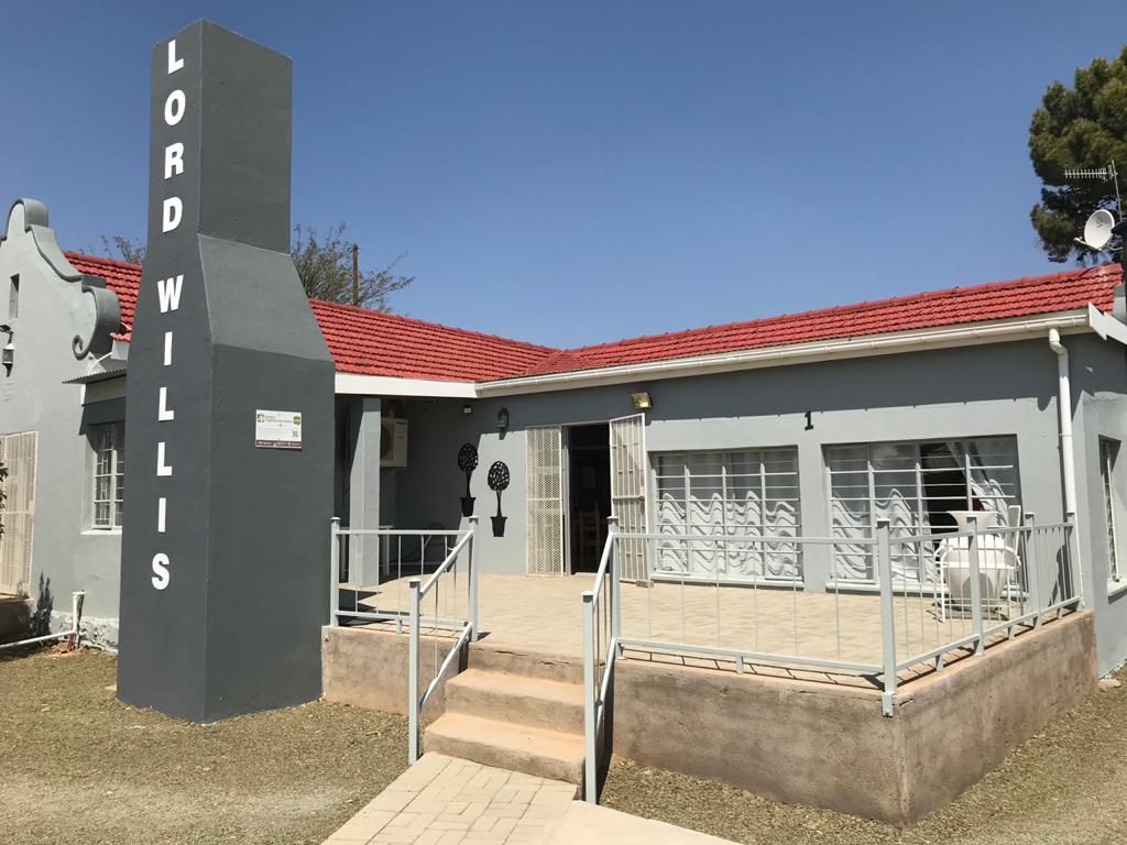 A front view of the building is presented, featuring a prominent gray structure with a red roof. Steps lead up to a spacious patio area, and a sign indicating 'Lord Willis' is visible on a tall chimney. Windows are arranged symmetrically across the facade.