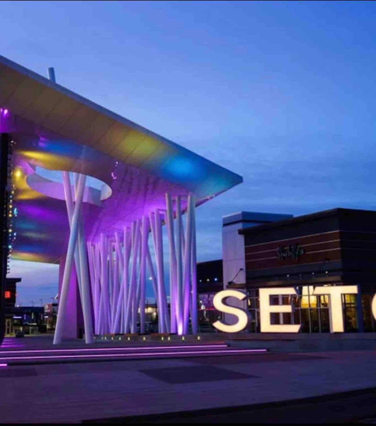 A vibrant architectural feature of Seton is showcased, with striking purple and blue lighting illuminating its modern design. The large 'SETON' sign stands prominently in the foreground, complemented by a contemporary building structure in the background.