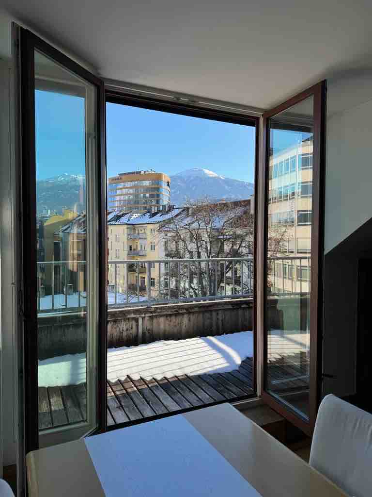 Large glass doors open to a sunny balcony, showcasing a view of distant mountains and surrounding buildings. A wooden deck is visible, highlighting the outdoor space. Bright light fills the room, indicating a spacious and airy atmosphere.