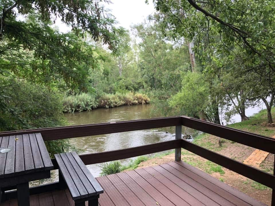 A wooden deck featuring a table and benches overlooks a serene river, bordered by lush greenery. The tranquil water reflects the surrounding trees, creating a peaceful atmosphere. The area is shaded, providing a comfortable spot for relaxation and enjoying nature.