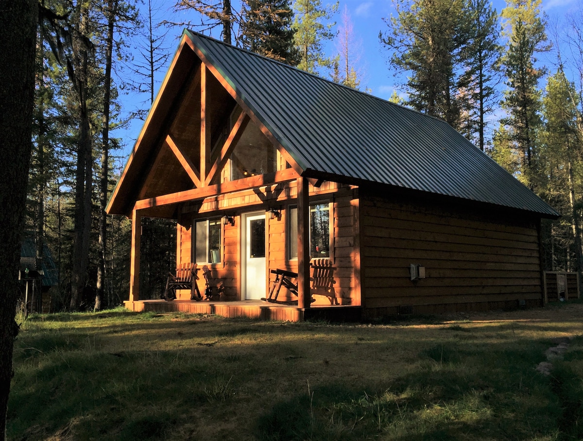 A handcrafted cabin features a warm wooden exterior with a sloped metal roof. The front porch is furnished with wooden rocking chairs and surrounded by lush trees. Sunlight casts gentle shadows, highlighting the cabin's inviting design.