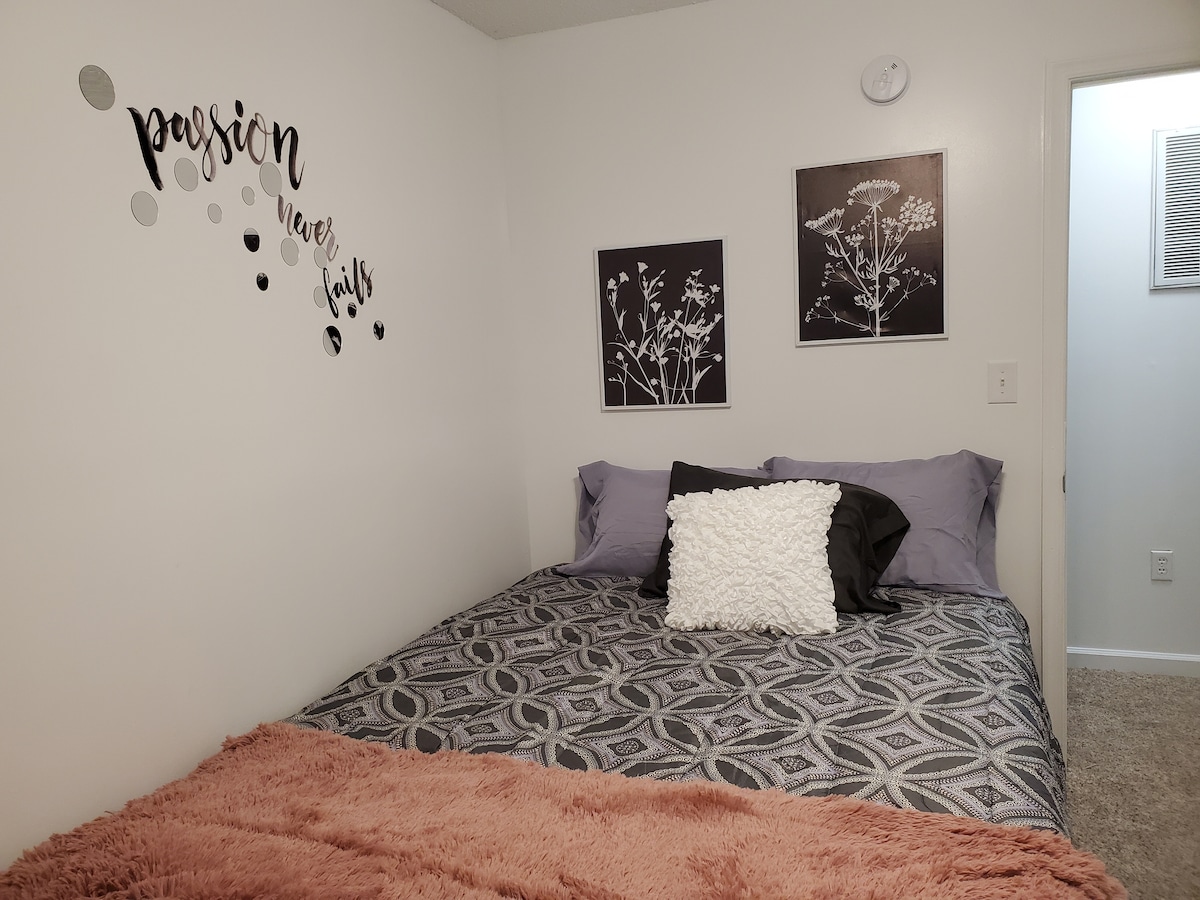 A private room features a large bed with a patterned blanket and decorative pillows. Wall art displaying floral designs complements a motivating phrase above the bed. A soft throw blanket lays across the foot of the bed. Light from a nearby window brightens the space.