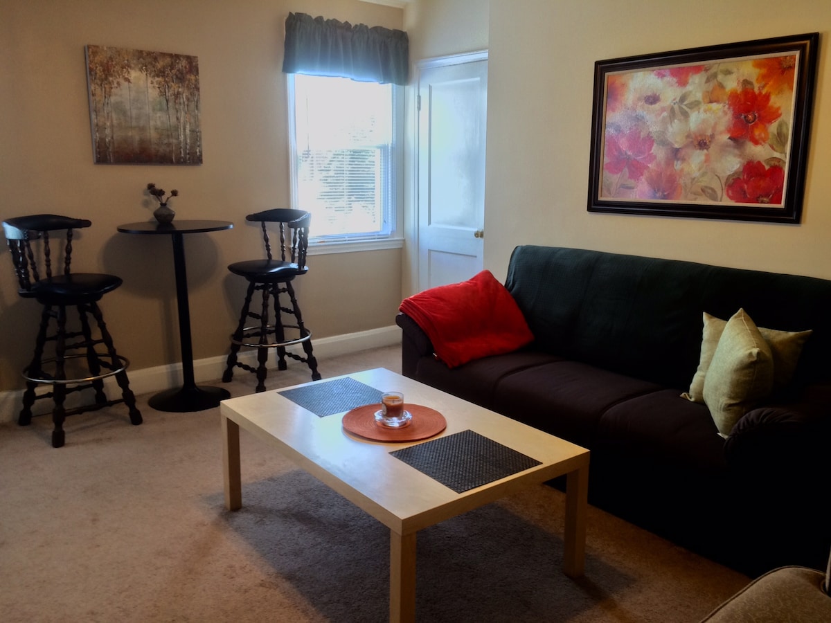 The living area features a casual seating arrangement with a dark sofa, a wooden coffee table, and two bar stools paired with a small round table. Natural light enters through the window beside the door, highlighting the pleasant wall art.