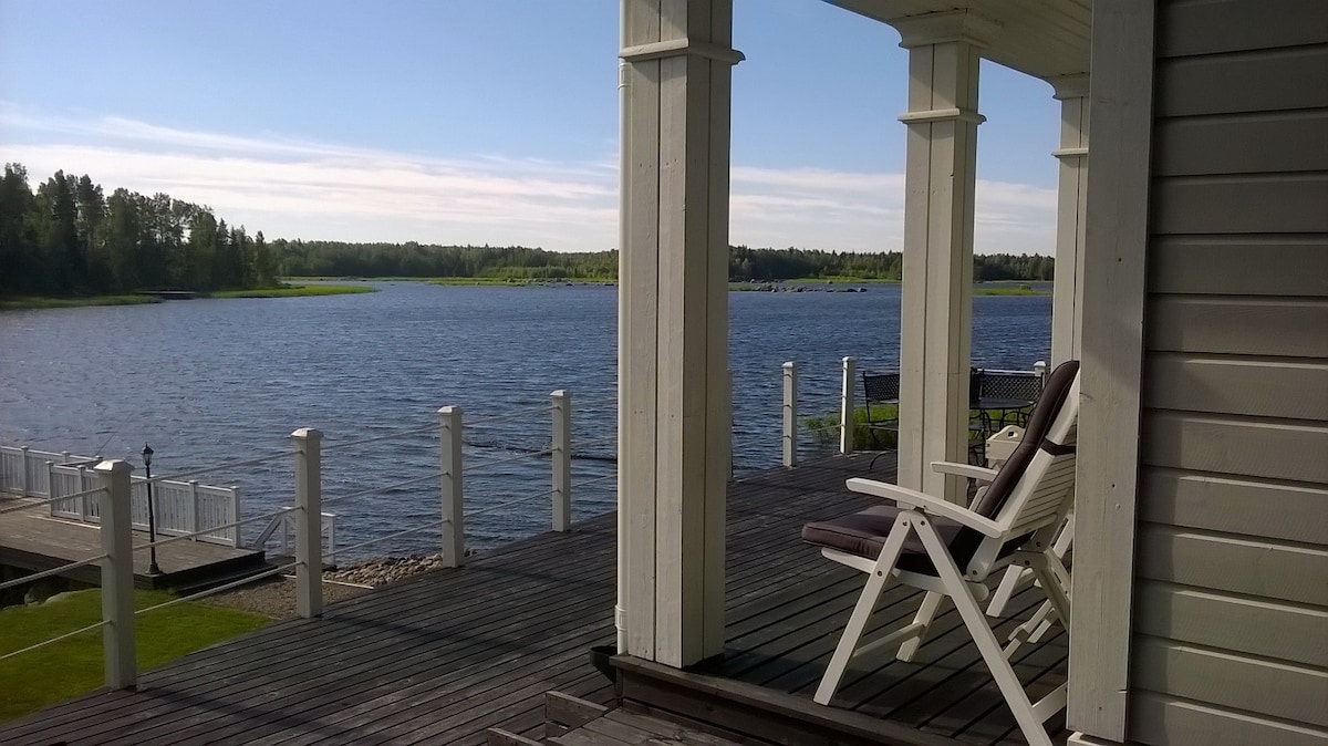A spacious deck is visible, featuring white pillars and overlooking a calm body of water. A single white chair is positioned on the deck, with green fields and trees seen in the distance under a clear blue sky.