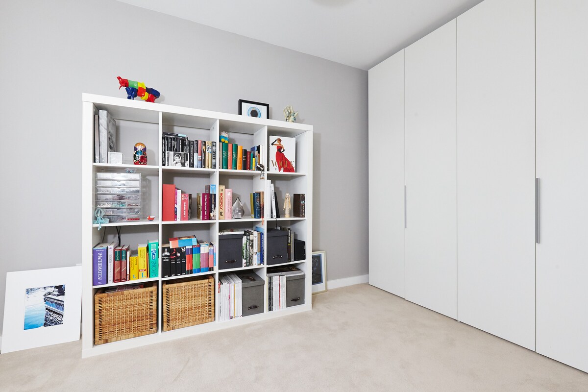 A light, airy room features a white shelving unit filled with colorful books and decorative items. A few woven baskets are placed on the lower shelves. To the right, a built-in wardrobe with white doors occupies the space, contributing to a tidy and organized look.
