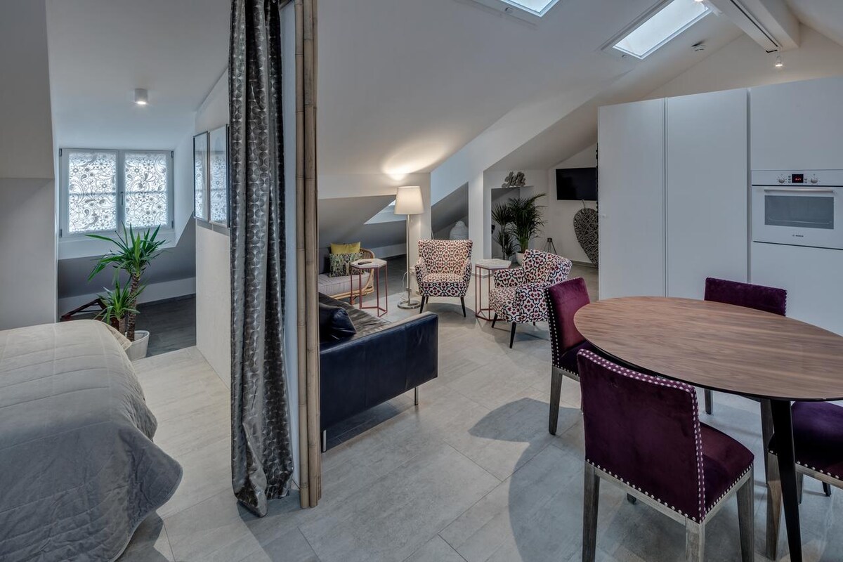 An open living area features a cozy sleeping space with a comfortable bed and two inviting chairs arranged around a small table. Natural light enters through skylights and windows, highlighting the modern decor. A flat-screen TV and kitchenette are visible in the background.