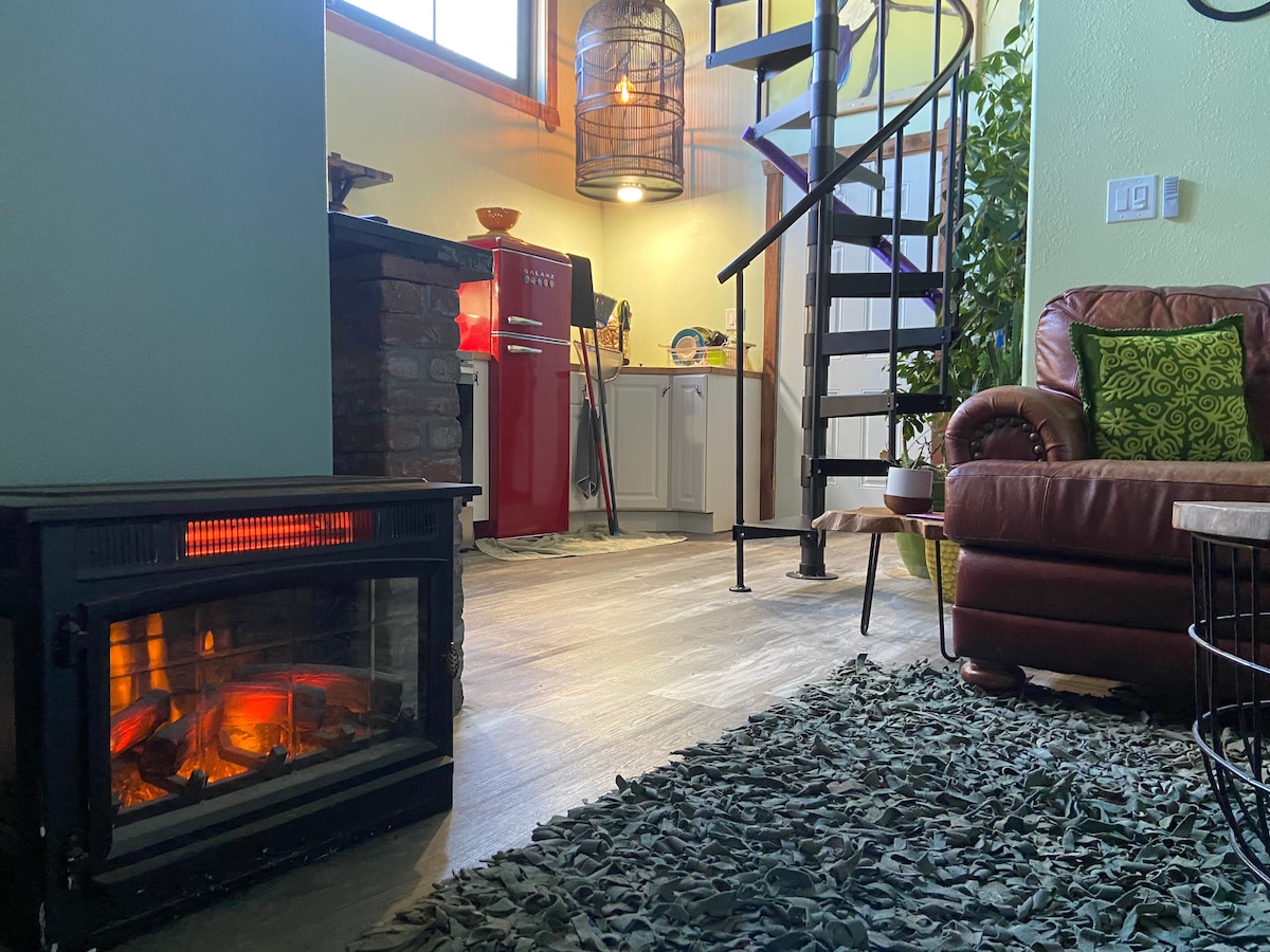An open living area features a spiral staircase, leading to the loft. A vintage red refrigerator stands alongside modern kitchen appliances. A cozy seating area is highlighted by a textured green decorative pillow. A fireplace adds warmth with its glowing embers, enhancing the inviting ambiance.