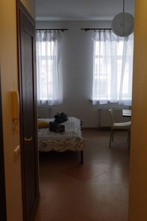 A bright, cozy room is seen through an open doorway, featuring a bed with floral bedding and neatly arranged towels. Large, sheer white curtains allow natural light to fill the space. A small table and chair are positioned nearby, enhancing the room's functionality.