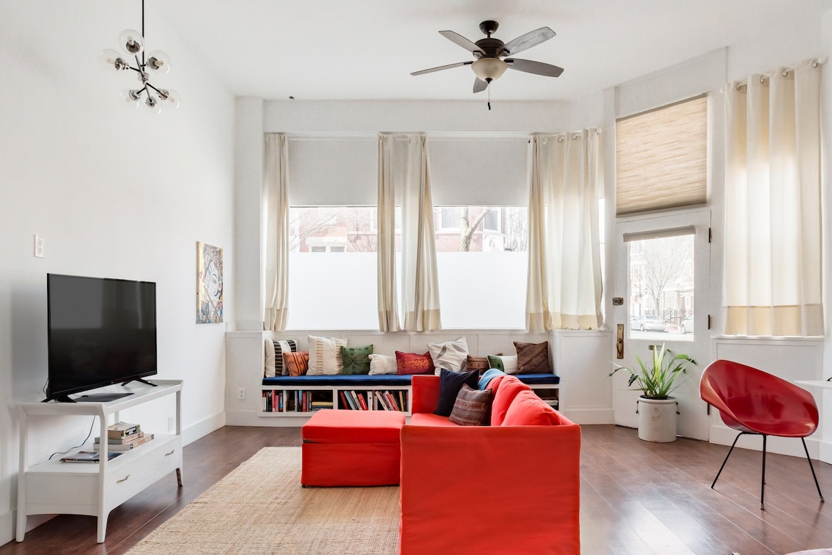A bright living area is highlighted by large windows draped with sheer curtains that allow natural light to fill the space. A red sectional sofa and an accent chair provide comfortable seating, paired with a coffee table and a television on a stand. Books are arranged on a shelf in the background.