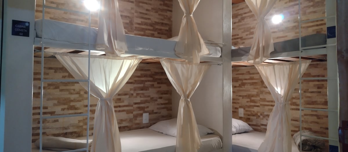 A shared sleeping area features multiple bunk beds, each with individual curtains for privacy. Soft lighting is provided by bedside lamps. The walls are adorned with textured natural stone, creating a cozy ambiance. Bedding is neatly arranged on each mattress, inviting relaxation.