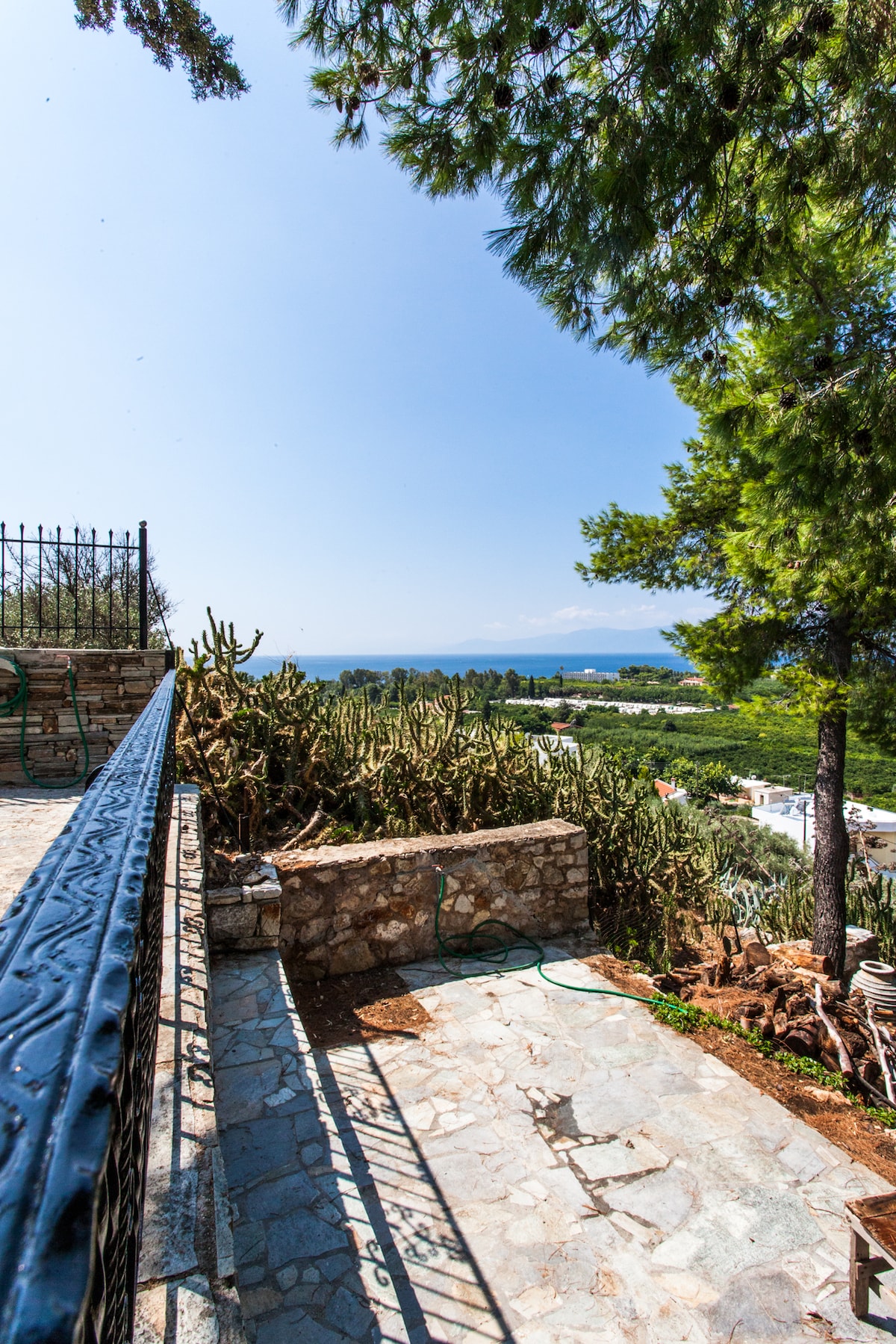 A sunlit terrace area is shown, featuring stone flooring and a metal railing. Cacti and greenery occupy the surrounding landscape, leading to a panoramic view of the sea in the distance, with a clear blue sky above.