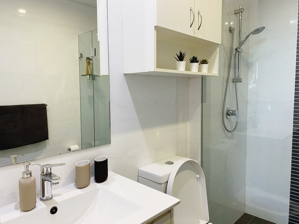 A modern en-suite bathroom features a sleek vanity with a sink and decorative candles. A shower area with a glass enclosure and a wall-mounted showerhead is visible. Minimalist shelving displays potted plants, while a dark towel is hung nearby.