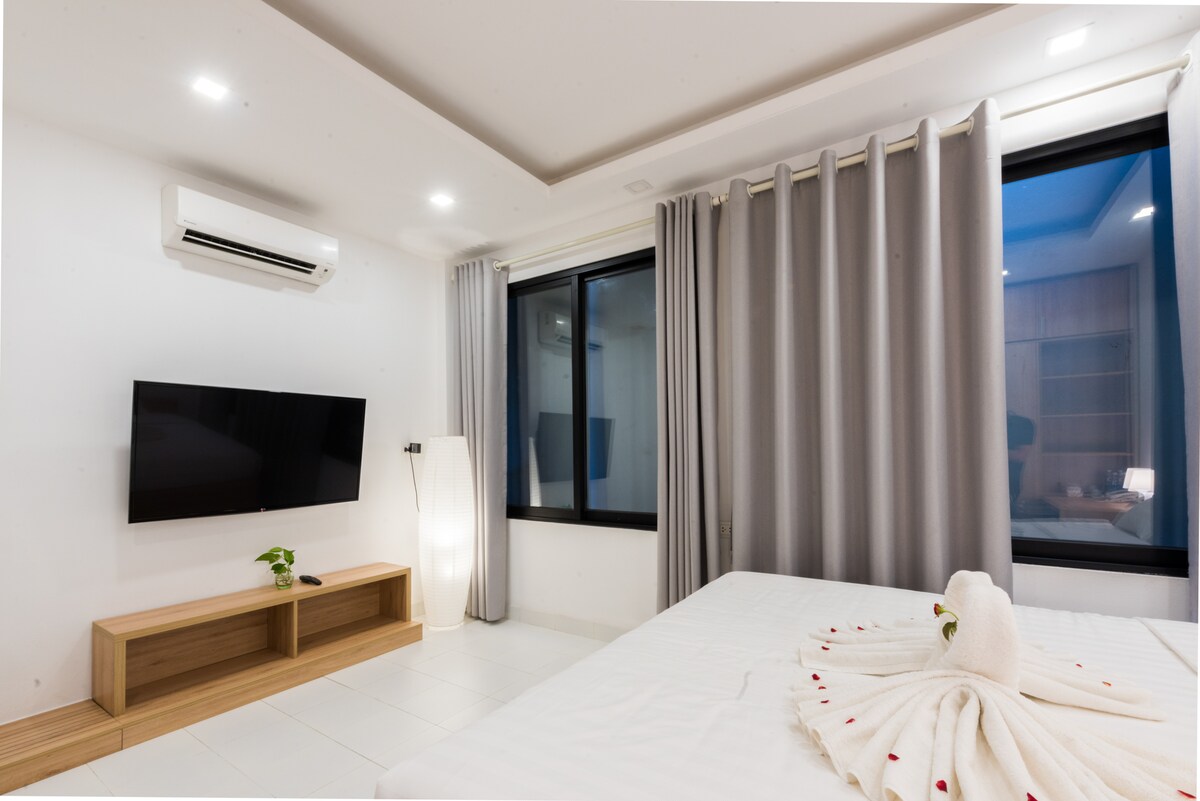 A modern en suite room is equipped with a flat 49’ wall-mounted TV and an air conditioning unit. Floor-to-ceiling windows draped with light gray curtains provide natural light. A minimalist wood console table sits beneath the TV, complementing the clean white walls and tiled floor.