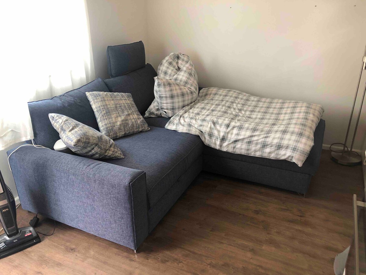 A cozy corner features a spacious sectional sofa adorned with patterned throw pillows and a lightweight plaid blanket. Natural light filters through a nearby window, illuminating the soft textures and inviting seating arrangement.