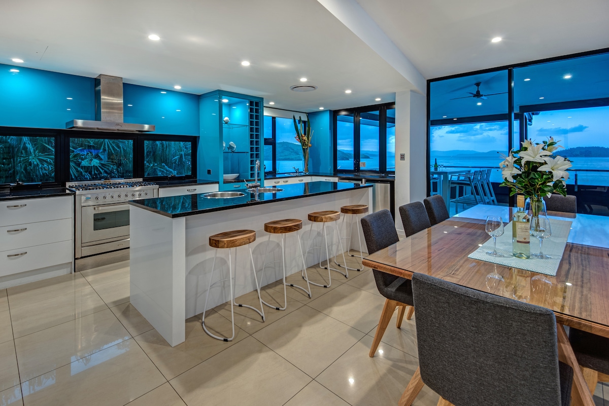 An open-plan kitchen and dining area features a modern design with blue accent walls and large windows showcasing ocean views. A spacious kitchen island with bar stools is complemented by top-of-the-line appliances, while an inviting dining table is set for meals with comfortable seating.