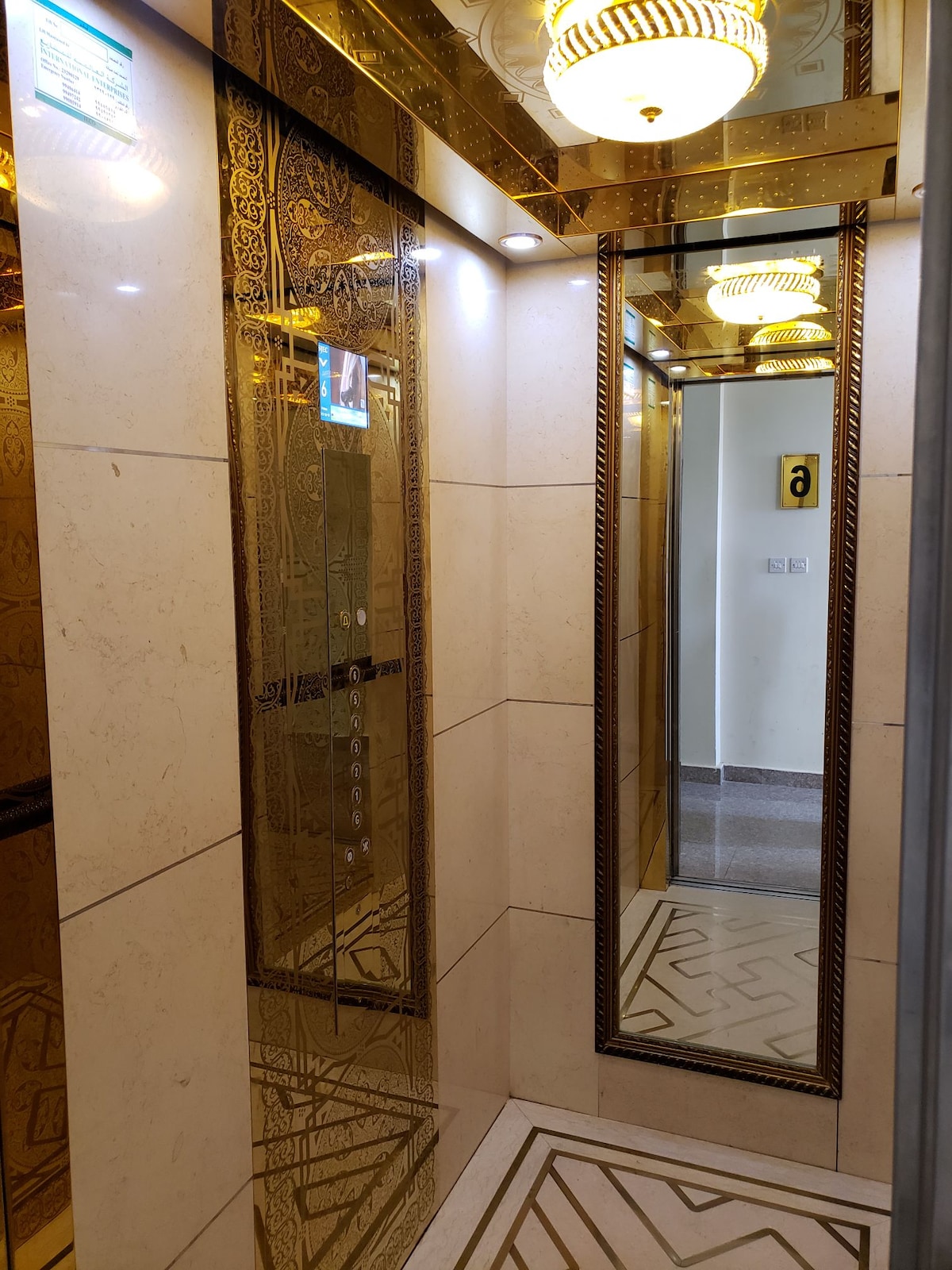 An elegantly decorated elevator interior features polished, reflective surfaces and intricate golden patterns. Stylish light fixtures illuminate the space, enhancing the design. The mirrored walls create a sense of openness, while the patterned floor adds a touch of sophistication.