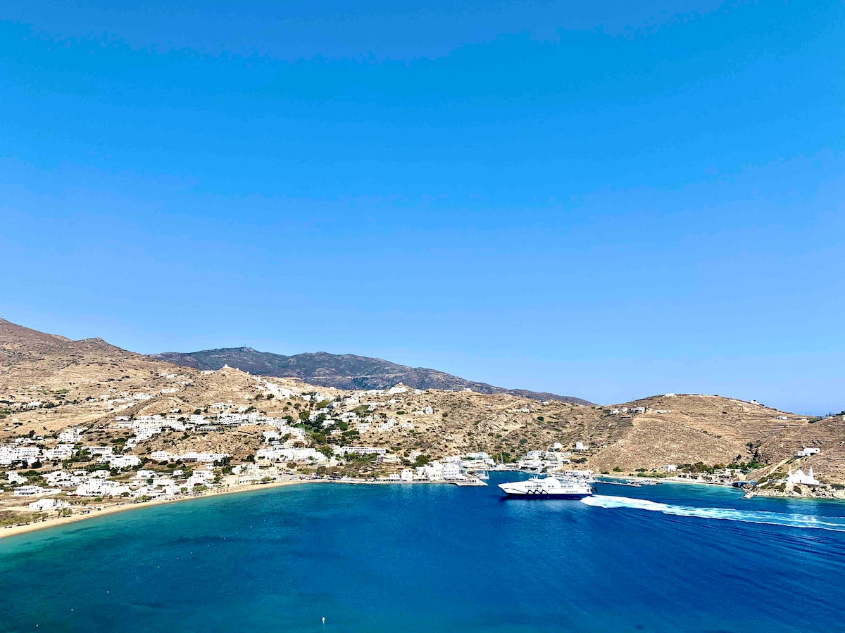 A panoramic view of a coastal landscape features clear blue waters gently lapping against sandy shores. Hills covered in white structures stretch into the distance under a bright blue sky, with a boat visible leaving a wake as it travels across the bay.