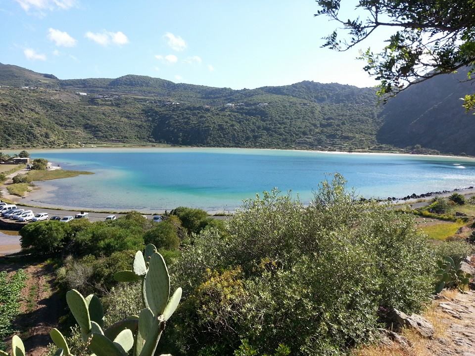 A serene view of a tranquil bay bordered by lush green hills and dotted with scattered vegetation. The calm waters reflect a variety of blues under the clear sky, while a small parking area is visible in the distance, framed by the natural landscape.