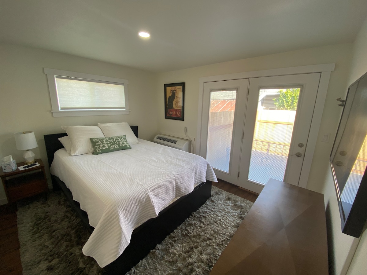 A well-lit bedroom features a spacious bed with a white quilt and decorative green pillow. A piece of artwork hangs on the wall, complementing the modern decor. Two glass doors open to a private outdoor area, while a bedside table with a lamp is positioned beside the bed.