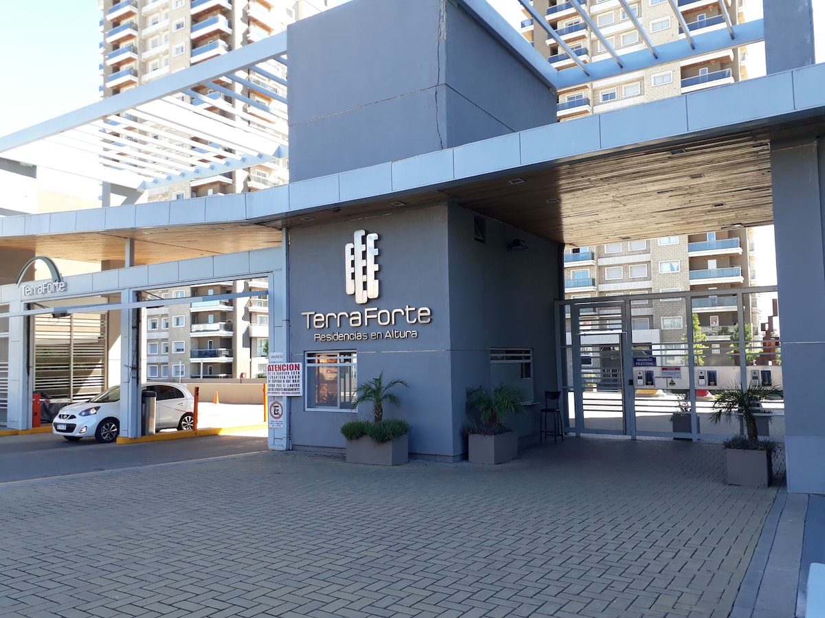 The entrance of the TernaForte complex is depicted, featuring a modern structure with a sign prominently displayed. Potted plants are arranged on either side of the entrance. The driveway leads to the building, surrounded by structured parking spaces and nearby high-rise buildings.