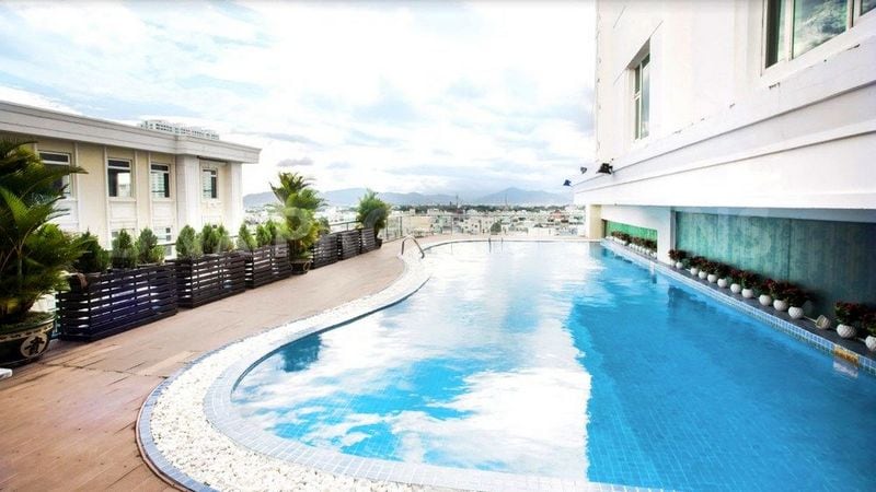 An inviting swimming pool is featured, positioned on an elevated deck surrounded by lush greenery. The water reflects the sky, providing a serene atmosphere. Stylish lounge areas can be seen along the edge, and the cityscape stretches into the distance.