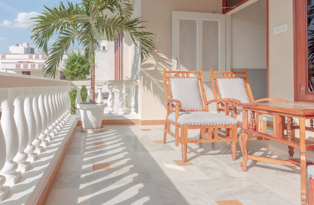 A serene balcony area is depicted with two wooden chairs facing a small table, complemented by a potted plant. Sunlight reflects off the marble flooring, enhancing the inviting atmosphere. The backdrop includes a glimpse of greenery and a clear sky, suggesting a peaceful outdoor space.