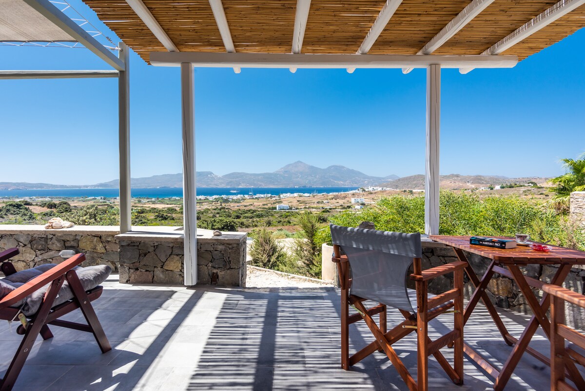 An outdoor terrace features wooden furniture, including a table and two chairs, offering expansive views of the coastal landscape. Sunlight illuminates the area beneath a bamboo roof, while the distant hills and water create a serene backdrop.