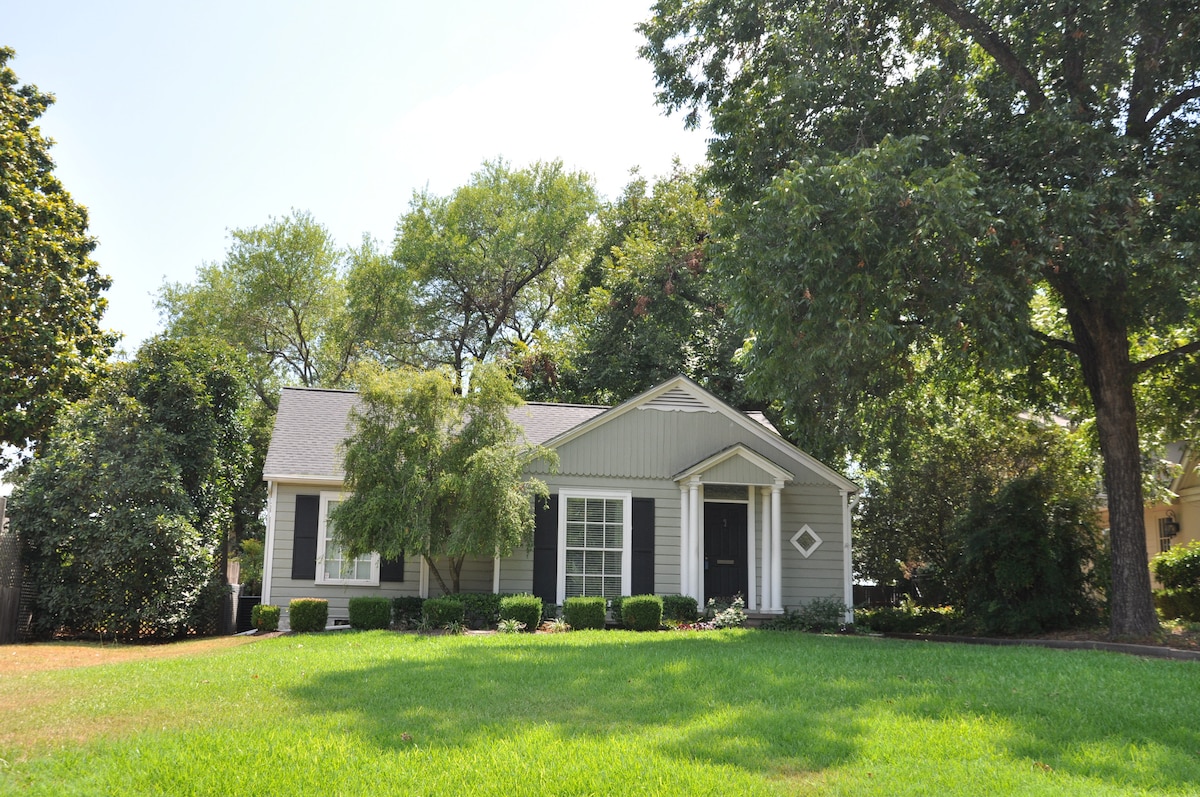 A charming cottage is set within a lush green lawn, surrounded by mature trees. The light gray exterior features a welcoming porch, large windows, and simple landscaping with neat hedges, providing a tranquil and inviting presence from the street.