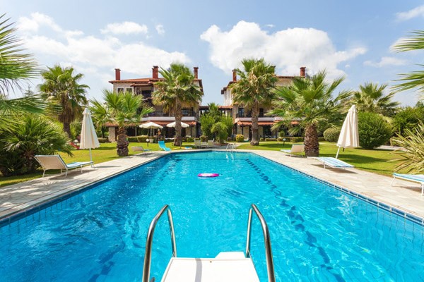 A large swimming pool stretches across the image, surrounded by palm trees and lush greenery. Lounge chairs and umbrellas are arranged around the pool deck, creating an inviting area for relaxation. Two building blocks are visible in the background, providing a glimpse of the accommodation.