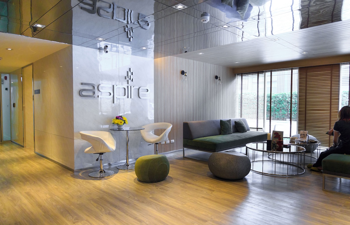 The lobby features a contemporary design with glossy surfaces and neutral tones. Two minimalistic chairs and a round table are situated beside a spacious seating area adorned with a plush sofa. Natural light filters through large windows, enhancing the inviting ambiance.