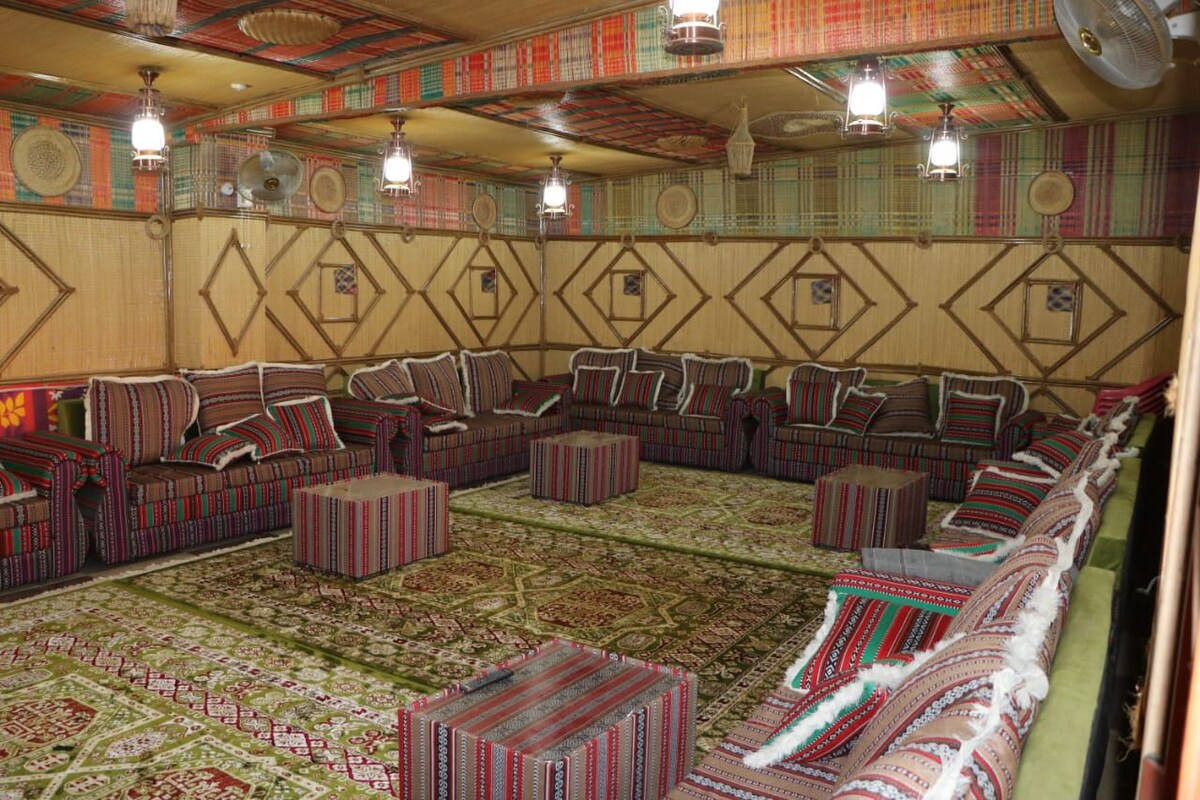 A spacious indoor gathering area features several patterned seating arrangements, including sofas and cube-shaped tables. The walls are adorned with vibrant textiles, and hanging light fixtures create a cozy atmosphere. A large decorative carpet covers the floor, providing a soft surface underneath.