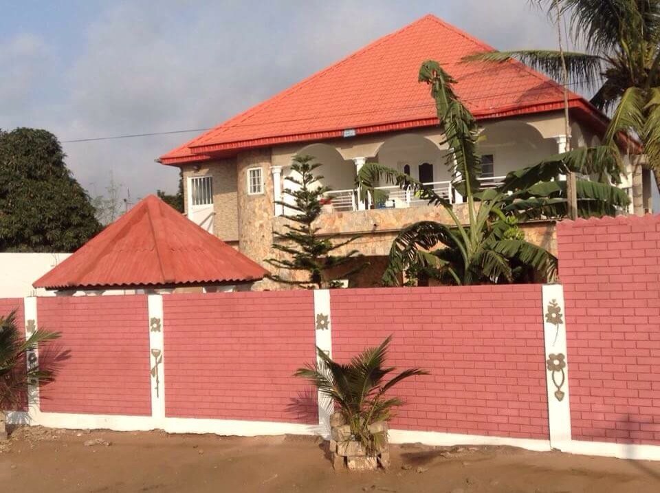 The exterior features a two-story house with a red-tiled roof and stone accents. A vibrant pink wall surrounds the property, providing privacy. Palm trees and other greenery can be seen in the foreground, complementing the inviting design of the home.