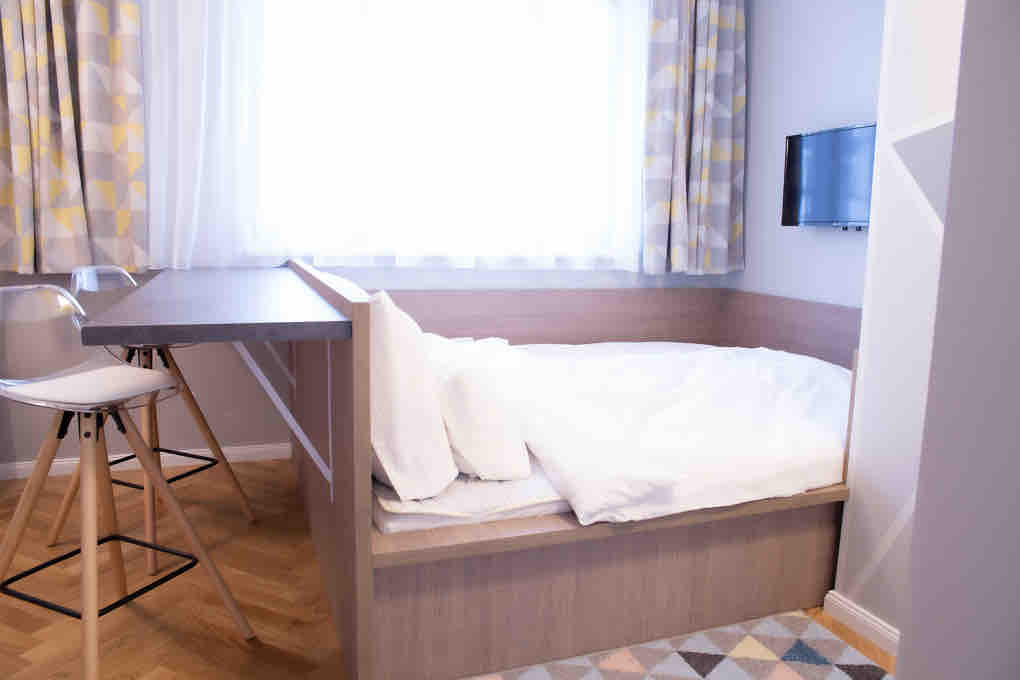 A cozy sleeping area features a spacious bed with white bedding positioned near a wall-mounted television. Light-colored curtains frame a large window, while a small dining table with two chairs is visible nearby, creating a functional and inviting space.